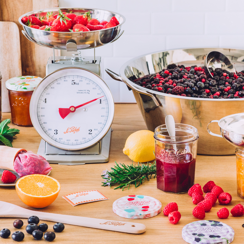 A styled kitchen scene with a vintage-style Le Parfait brand analog scale holding a bowl of fresh strawberries, surrounded by berries, Jam jars of preserves, citrus, and small pastries on a wooden countertop.