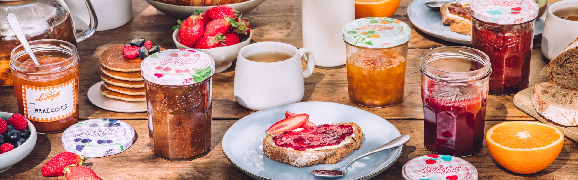 A rustic breakfast table set with Le Parfait jam jars, fresh fruit, and jam on toast
