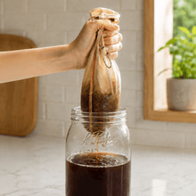 Load image into Gallery viewer, A hand holds a cloth bag filled with dark coffee grounds above a clear glass Le Parfait Screw Top 2L jar, with coffee dripping into the jar making cold brew in a bright kitchen setting.