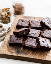Load image into Gallery viewer, Chocolate brownies cut into squares on a wood board with chopped walnuts and a jar of cocoa.