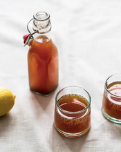 Load image into Gallery viewer, Swing-top bottle and two glasses of amber kombucha beside a fresh lemon on a linen tablecloth.