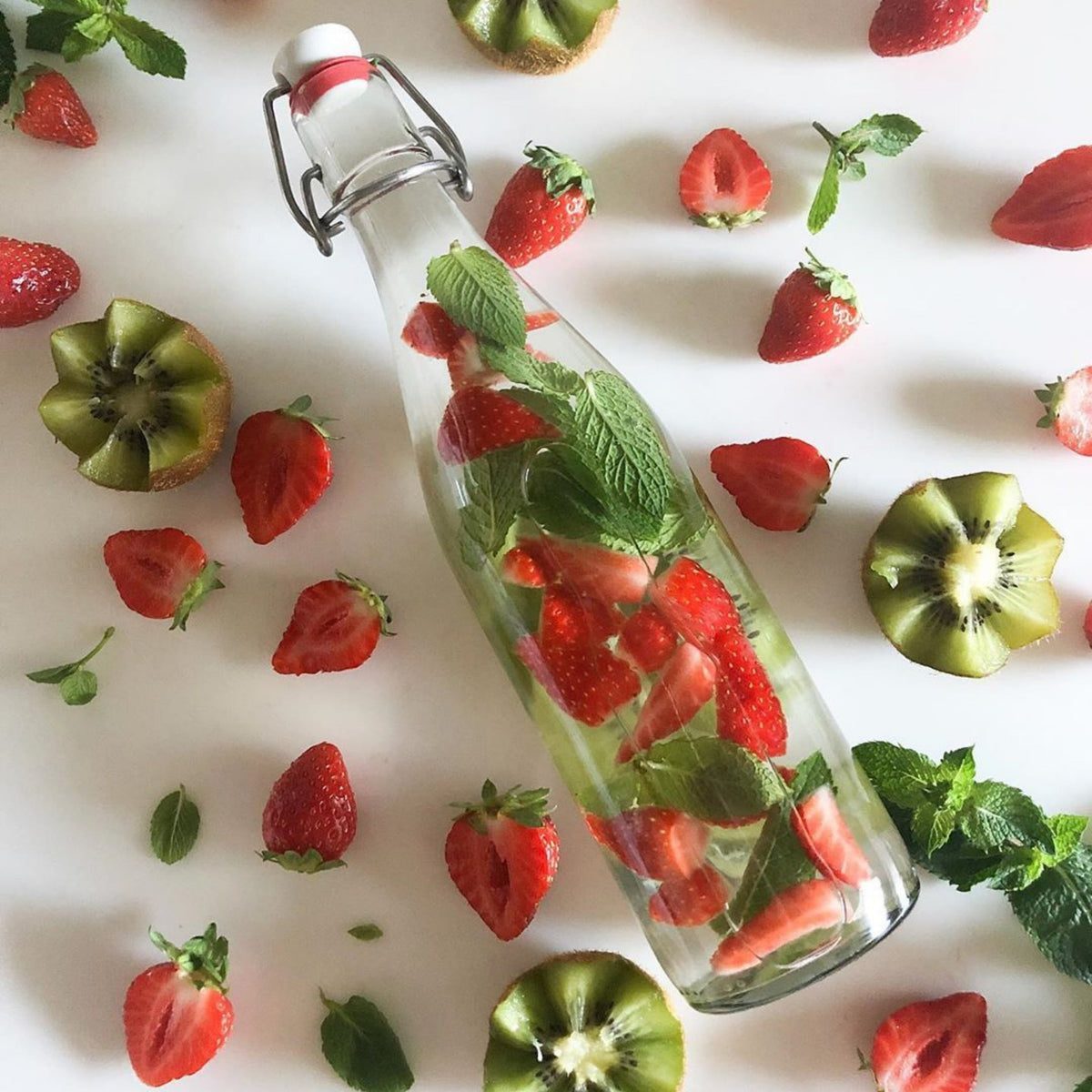 Top-down Le Parfait Swing Top Bottle of strawberry-mint infused water beside halved strawberries, mint leaves, and sliced kiwi.