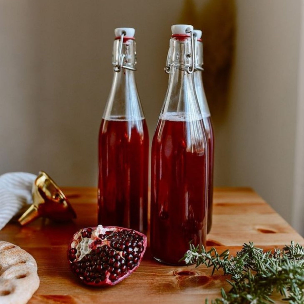 Two Le Parfait Swing Top Bottles of pomegranate cordial beside a halved pomegranate, rosemary, and a brass funnel on wood.