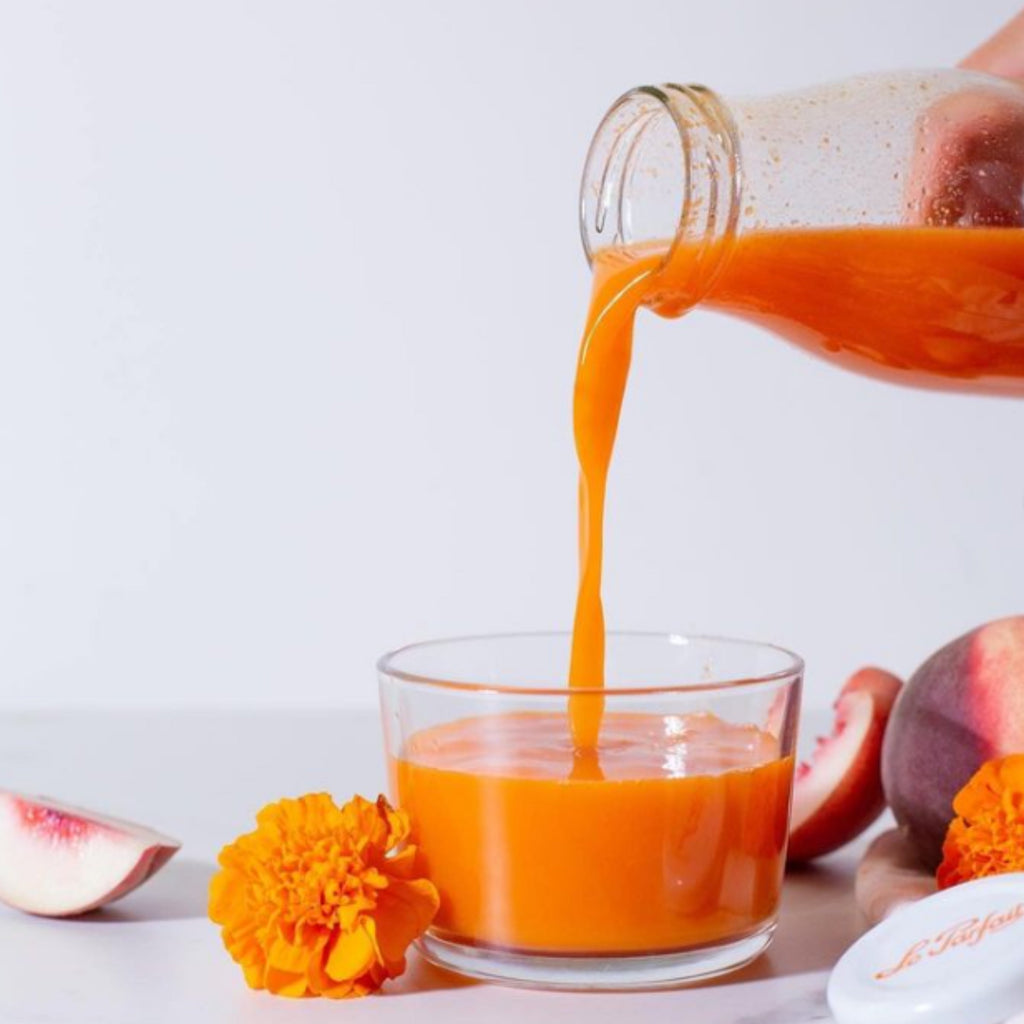 Pouring carrot-peach juice from a Le Parfait Milk Bottle into a glass cup beside peach slices and a marigold flower.