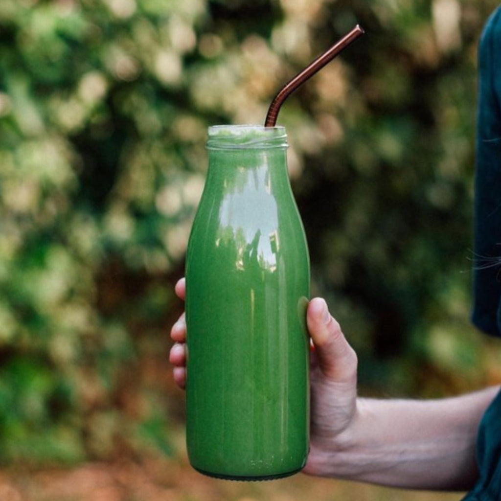 Hand holding a Le Parfait Milk Bottle filled with green smoothie outdoors with a copper straw.