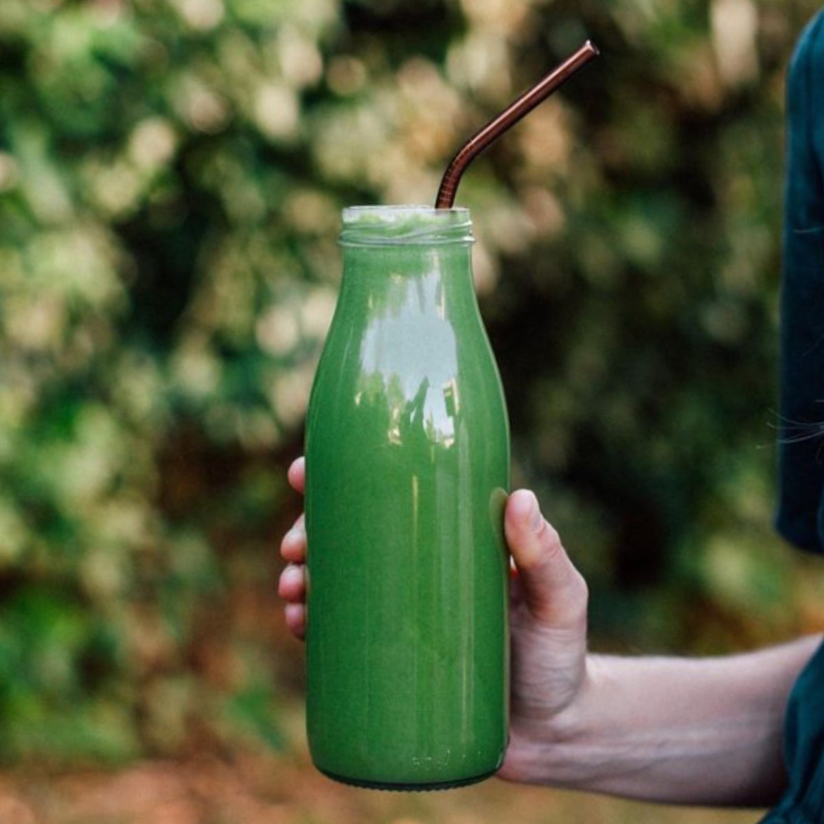 Hand holding a Le Parfait Milk Bottle filled with green smoothie outdoors with a copper straw.