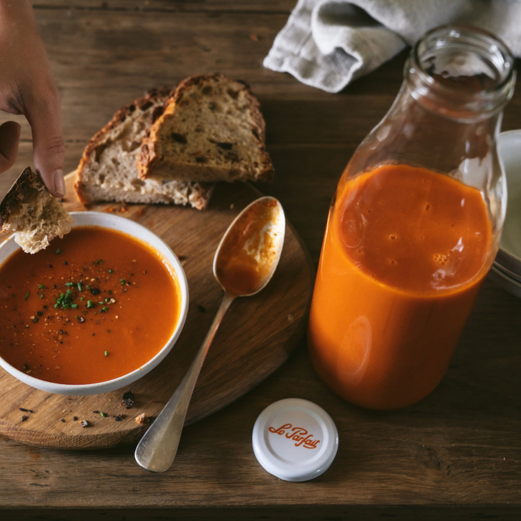 Le Parfait Milk Bottle filled with creamy tomato soup beside a served bowl with bread on a rustic wooden table.