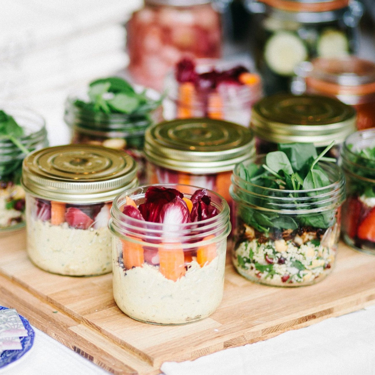 Layered Le Parfait Familia Wiss terrines packed with hummus, vegetables, and quinoa as make-ahead salads on a wooden serving board.