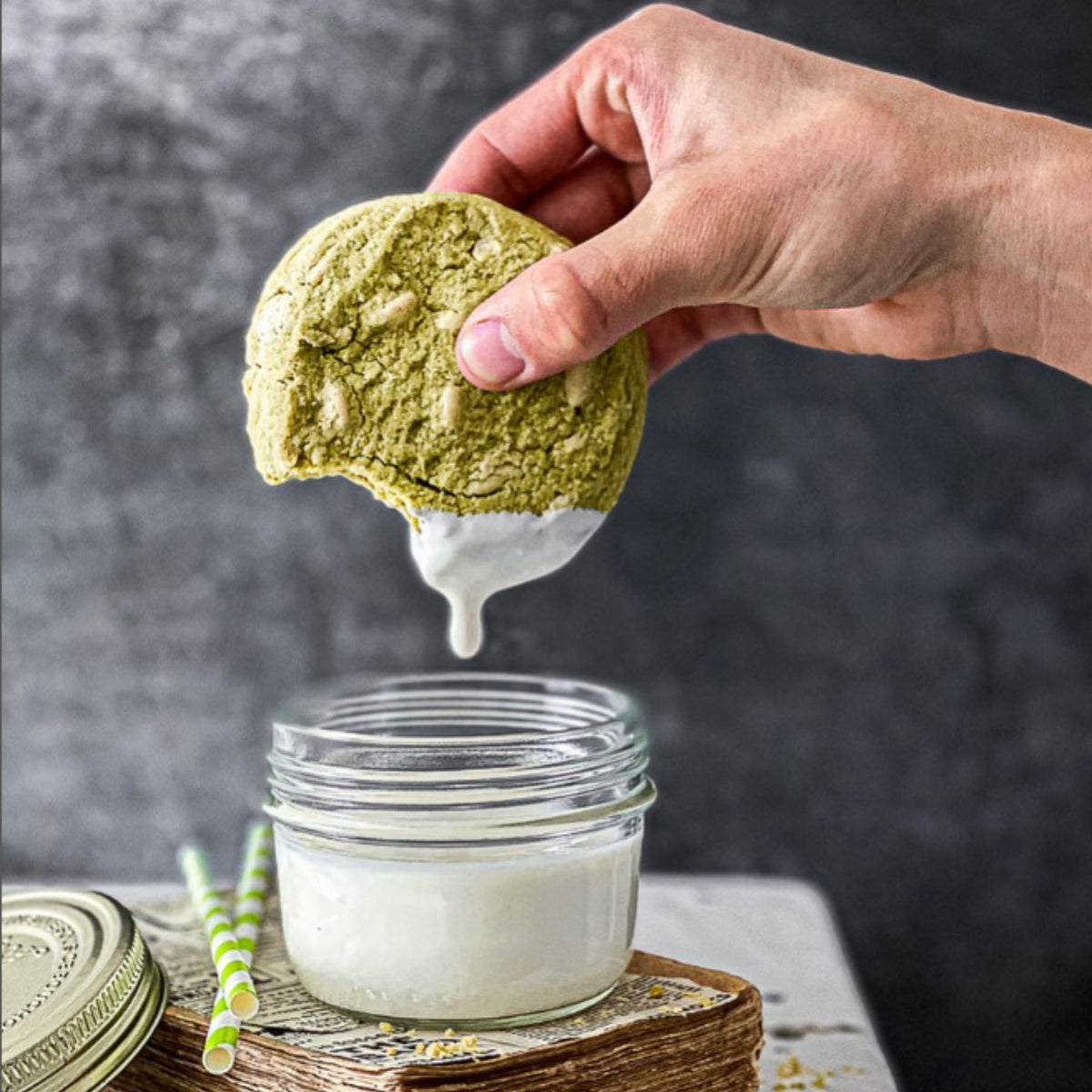Hand dipping a matcha cookie into a small Le Parfait Familia Wiss terrine of milk on a wooden board with green-striped paper straws.