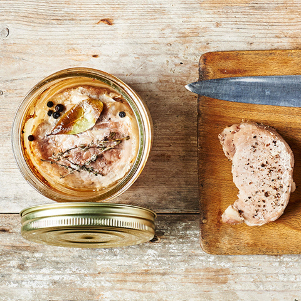 Top-down Le Parfait Familia Wiss terrine of pork rillettes with bay leaf, thyme, and peppercorns, beside raw pork on a cutting board.