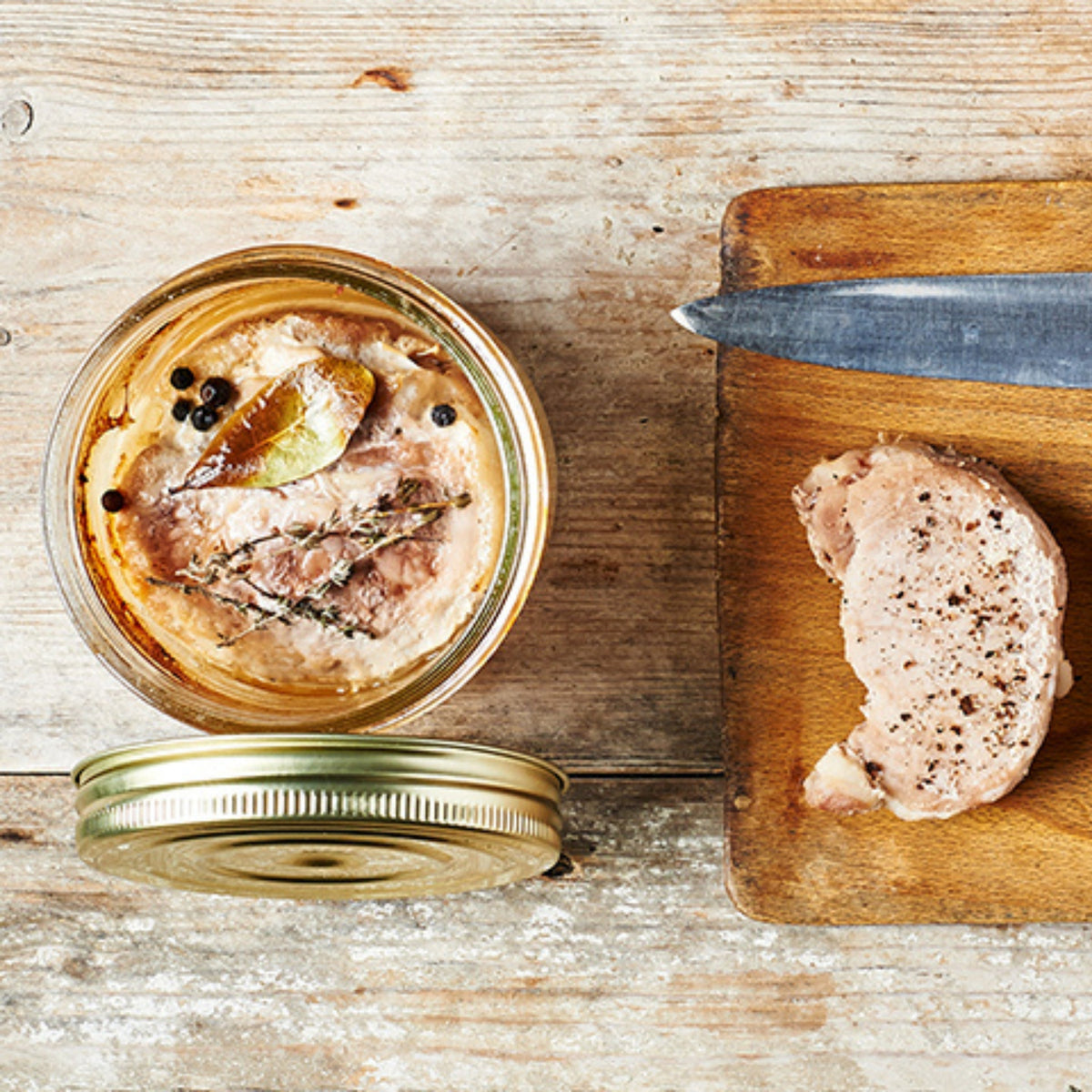 Top-down Le Parfait Familia Wiss terrine of pork rillettes with bay leaf, thyme, and peppercorns, beside raw pork on a cutting board.