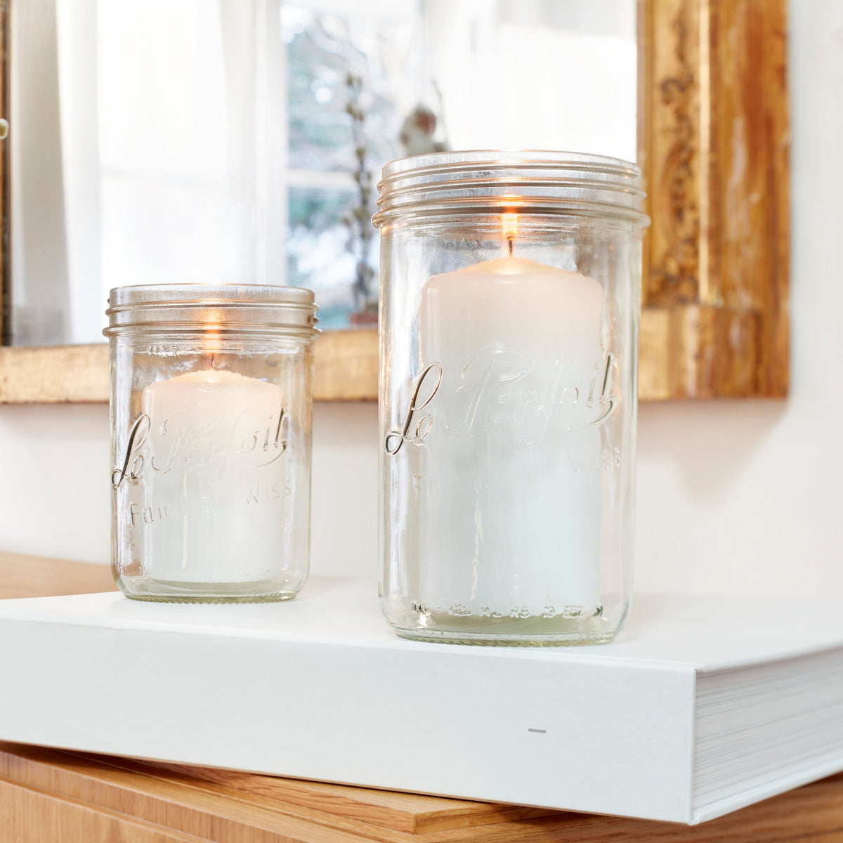 Two Le Parfait Familia Wiss terrines repurposed as candle holders with lit white pillar candles on a console table.