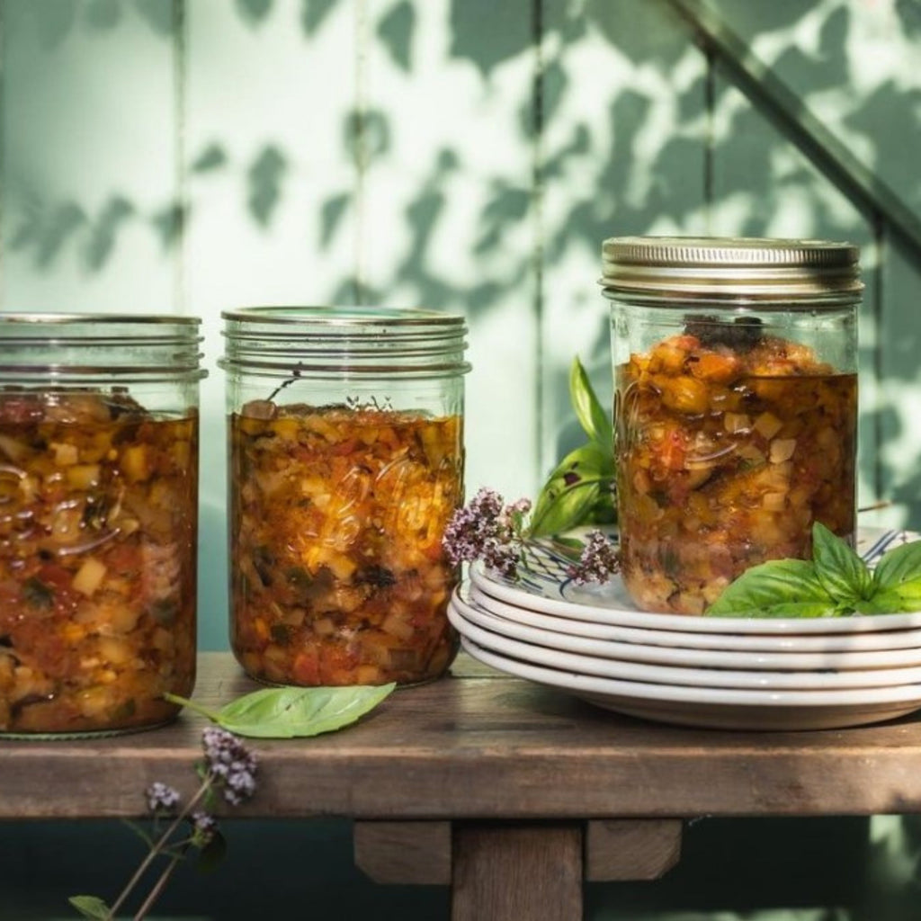 Three Le Parfait Familia Wiss terrines filled with ratatouille on a sunlit wooden bench with stacked plates and fresh basil.