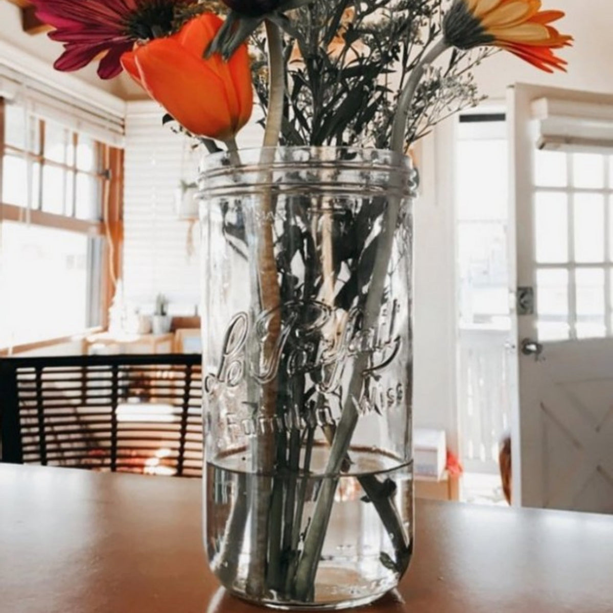 Tall Le Parfait Familia Wiss terrine repurposed as a vase with mixed gerbera daisies and a tulip on a sunny dining table.