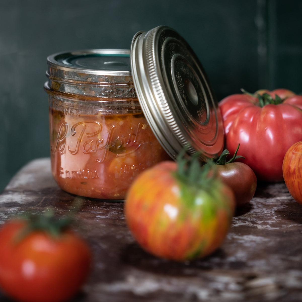 Le Parfait Familia Wiss terrine of homemade tomato sauce beside a cluster of heirloom tomatoes on a rustic stone surface.