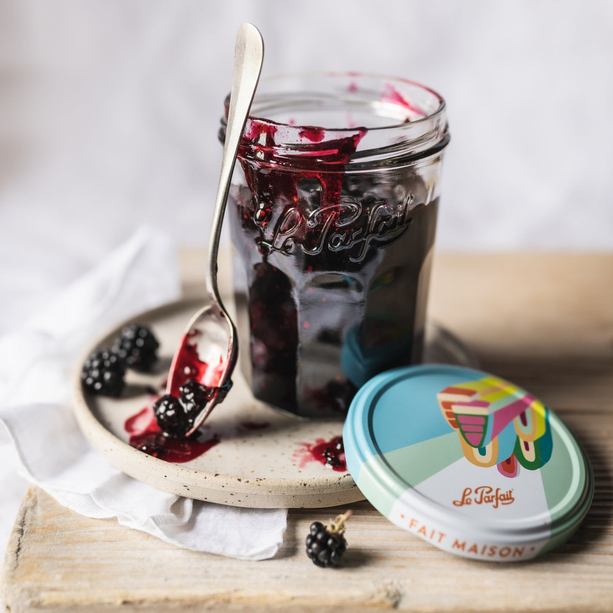 Open Le Parfait Jam Jar of blackberry preserves with silver spoon on a speckled plate, alongside fresh blackberries and a patterned 'fait MAISON' lid.