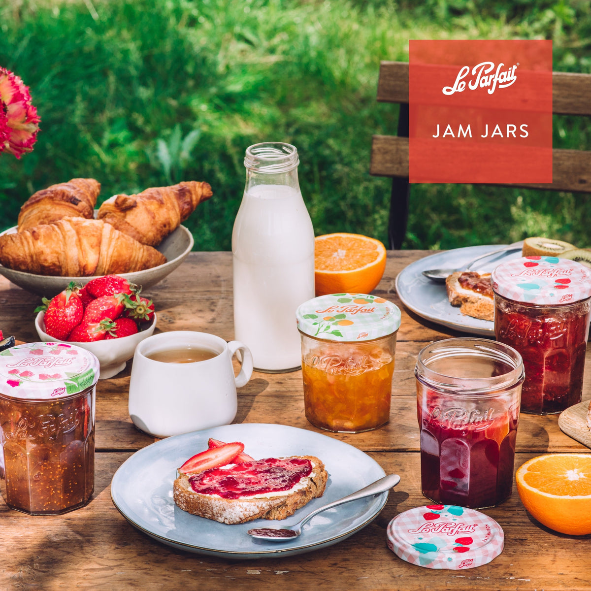 Outdoor breakfast spread with Le Parfait Jam Jars of strawberry, apricot, and raspberry preserves beside croissants, milk, tea, and sliced bread.