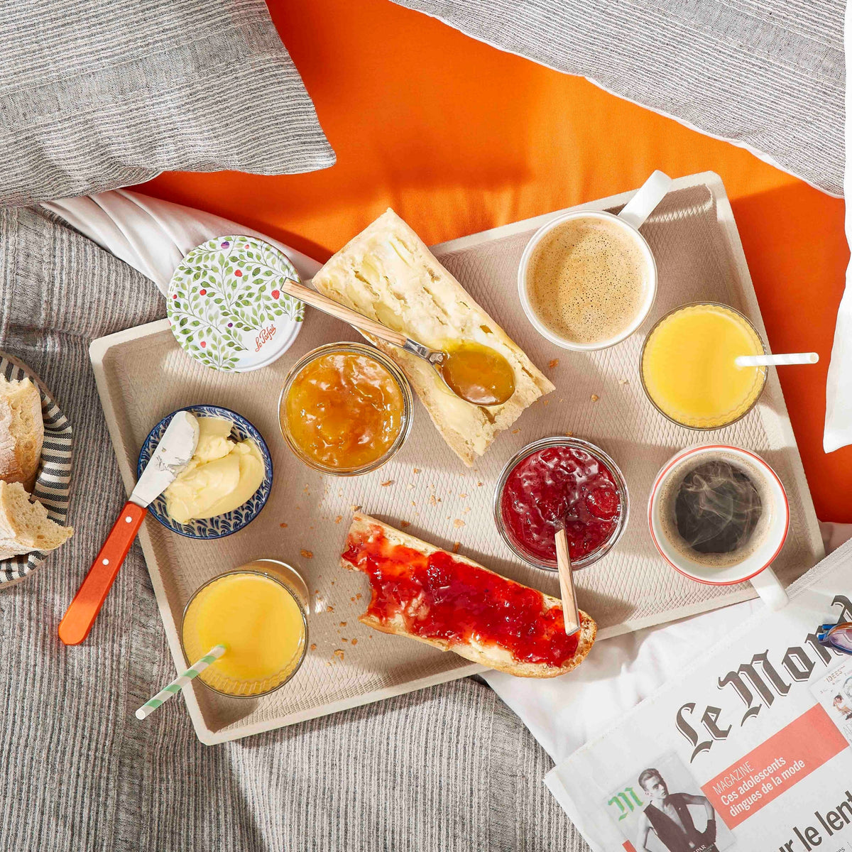 Top-down tray with open Le Parfait Jam Jars of orange marmalade and red preserves beside butter, baguette, juice, coffee, and Le Monde newspaper.