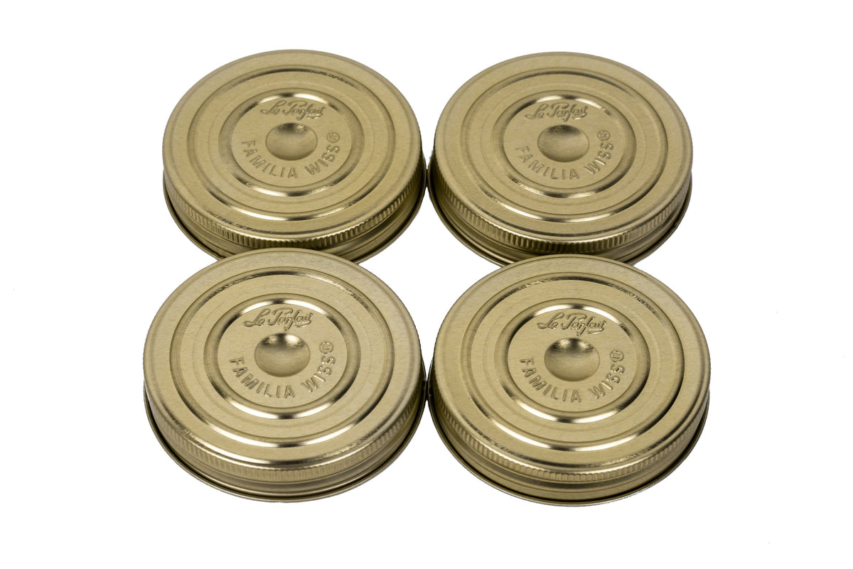 Four gold Familia Wiss metal canning lids by Le Parfait with embossed logo, angled product shot.