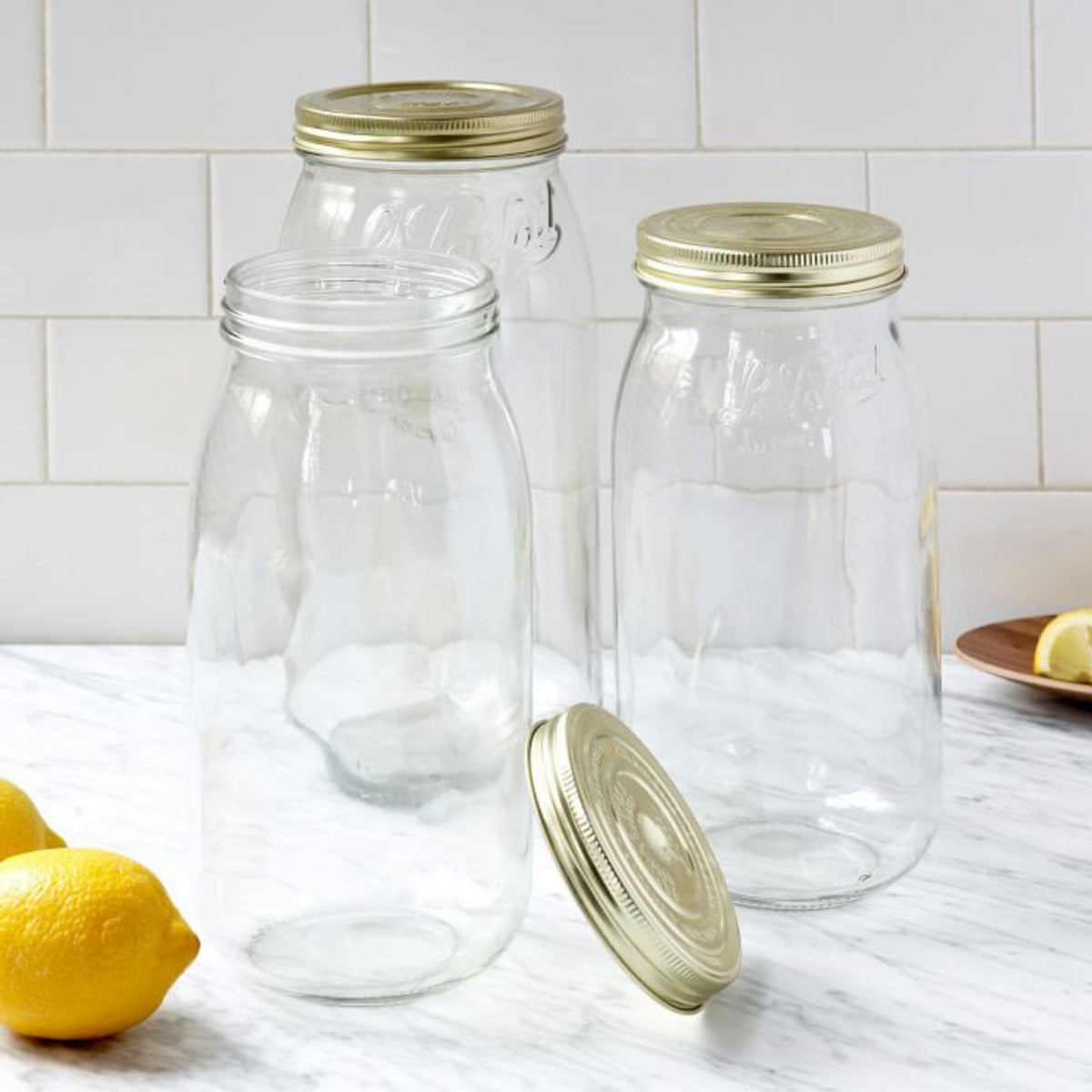 Three empty Le Parfait Super jars with gold metal canning lids on a white marble counter beside fresh lemons, one jar open with its lid alongside.