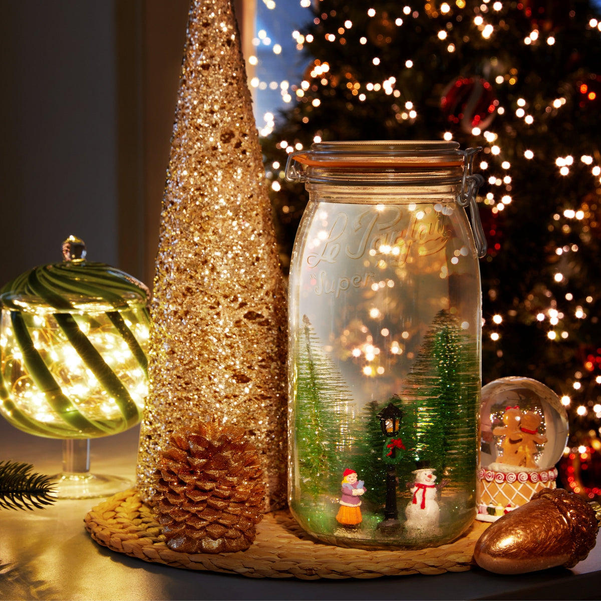 Le Parfait Super Jar styled as a holiday snow globe with miniature Christmas trees and figurines beside a lit Christmas display.