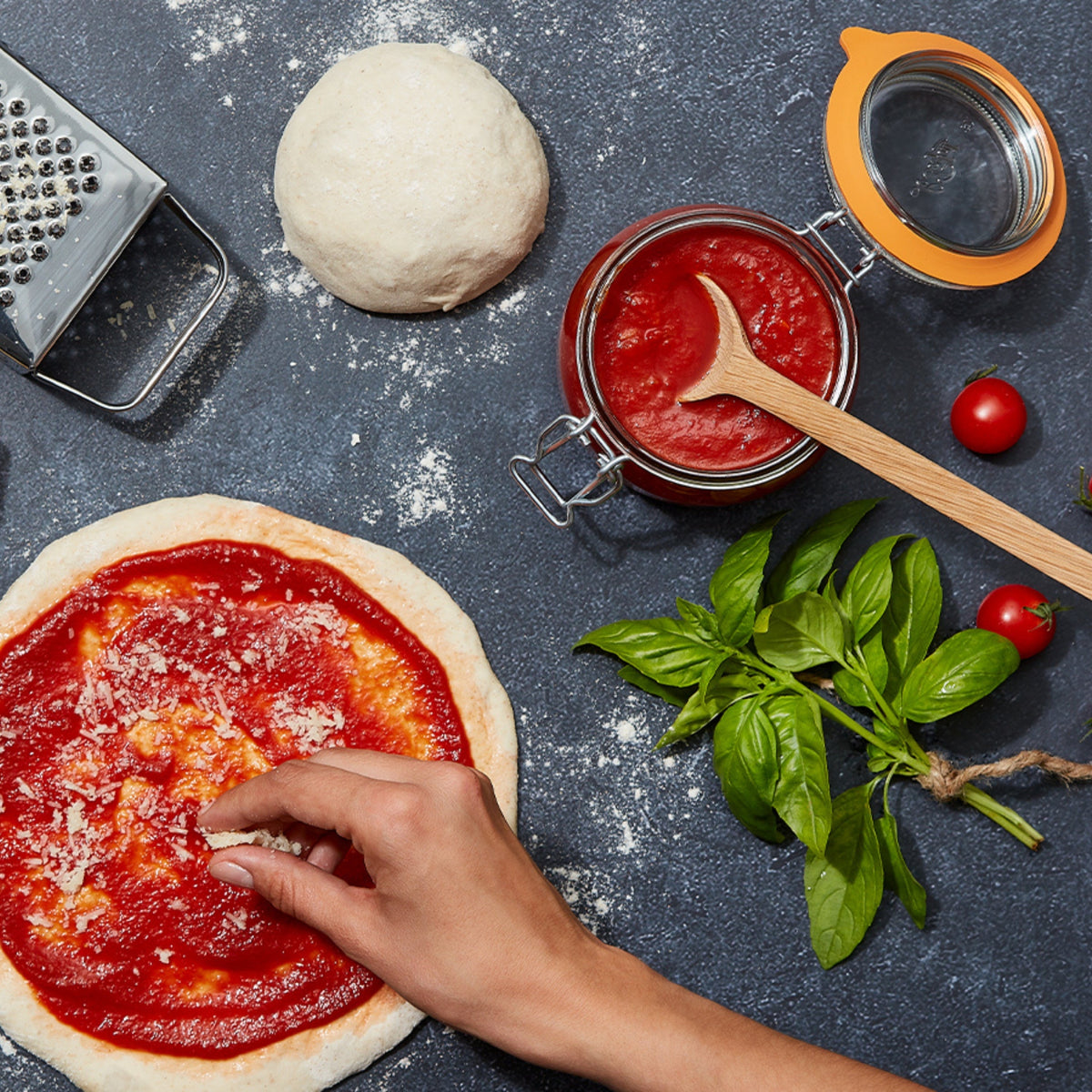 Overhead pizza prep scene with a Le Parfait Super Jar of tomato sauce, fresh dough, basil, cherry tomatoes, and a parmesan grater.