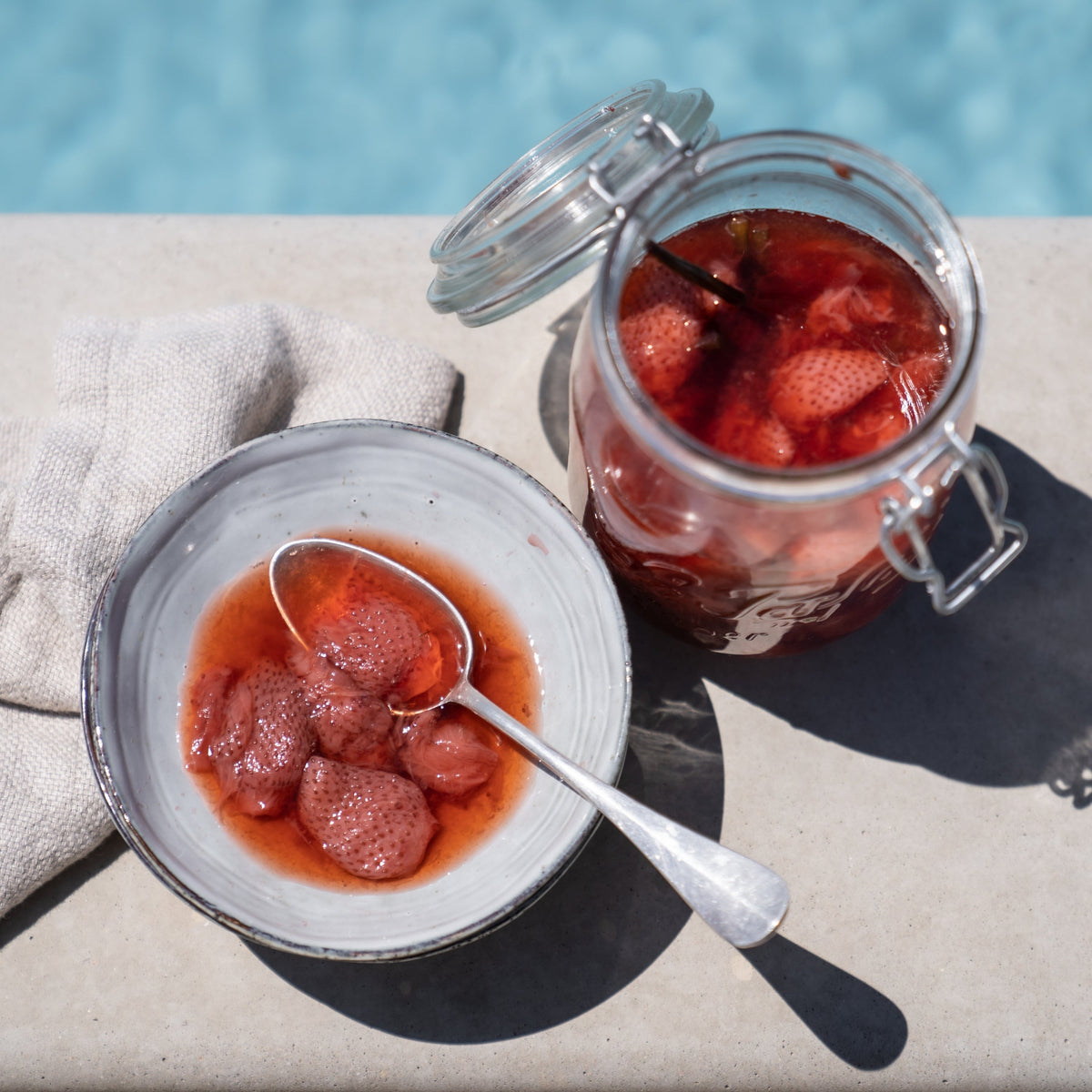Open Le Parfait Super Jar of preserved strawberries beside a bowl and silver spoon, on a sunlit poolside table.