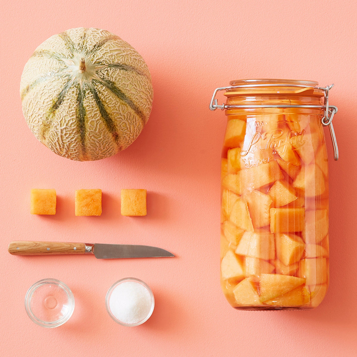 Cantaloupe melon being preserved in a tall Le Parfait Super Jar, with melon cubes, a paring knife, and sugar arranged on a coral background.