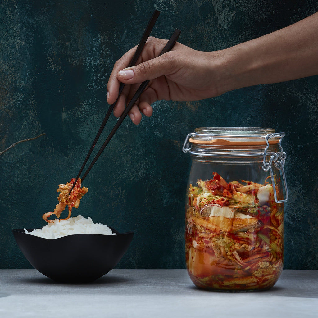Hand lifting fermented kimchi from a large Le Parfait Super Jar onto a bowl of steamed rice with chopsticks.
