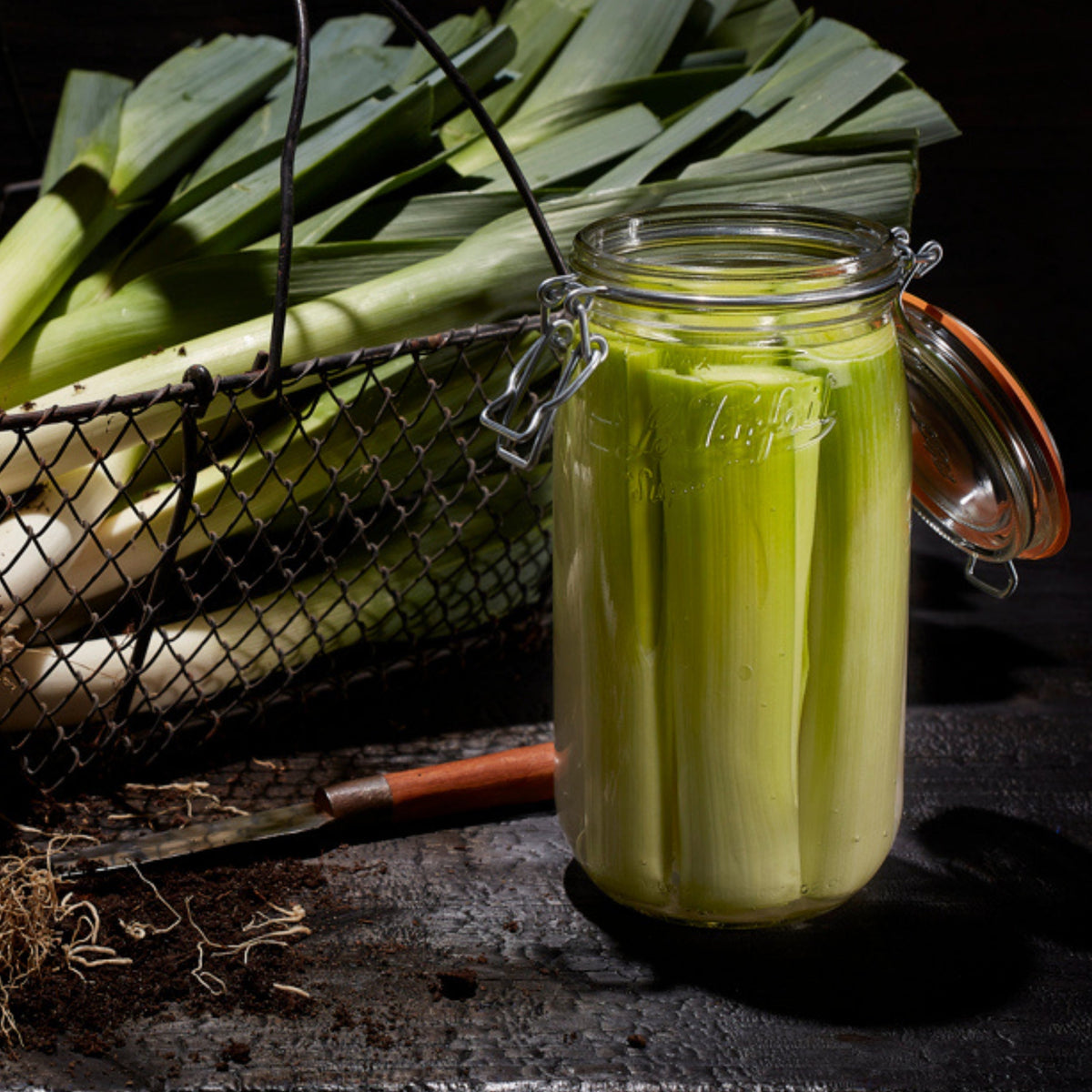 Tall Le Parfait Super Jar packed with fermenting leeks beside a wire basket of fresh leeks and a paring knife on a dark wood surface.