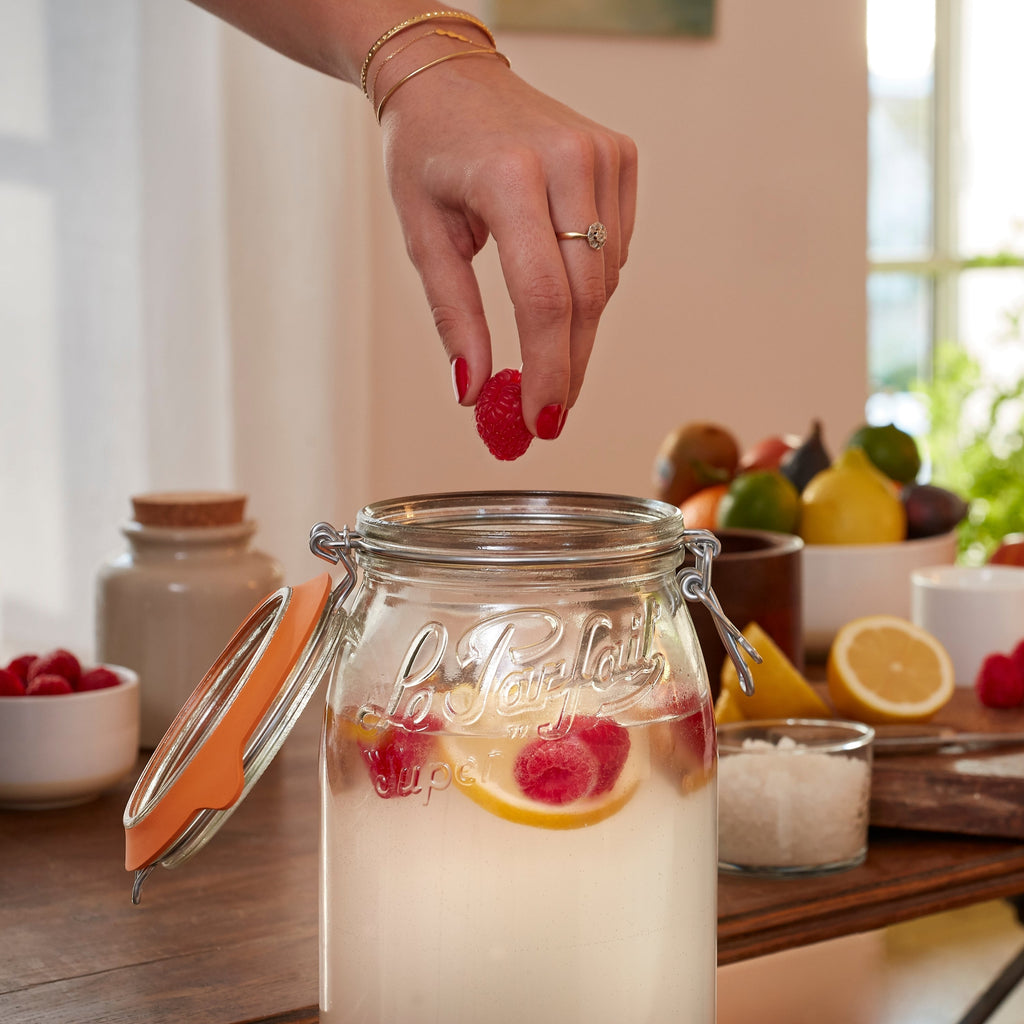 Hand dropping a raspberry into an open Le Parfait Super Jar of homemade fruit-infused beverage, with citrus and salt on the counter.