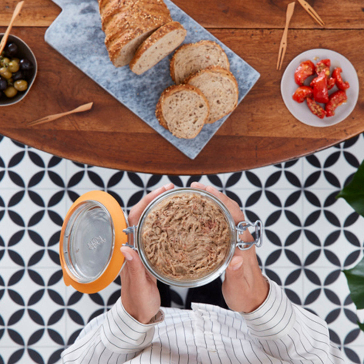 Overhead view of hands holding an open Le Parfait Super Terrine of pork rillettes over a tiled floor with bread, olives, and pickled peppers.