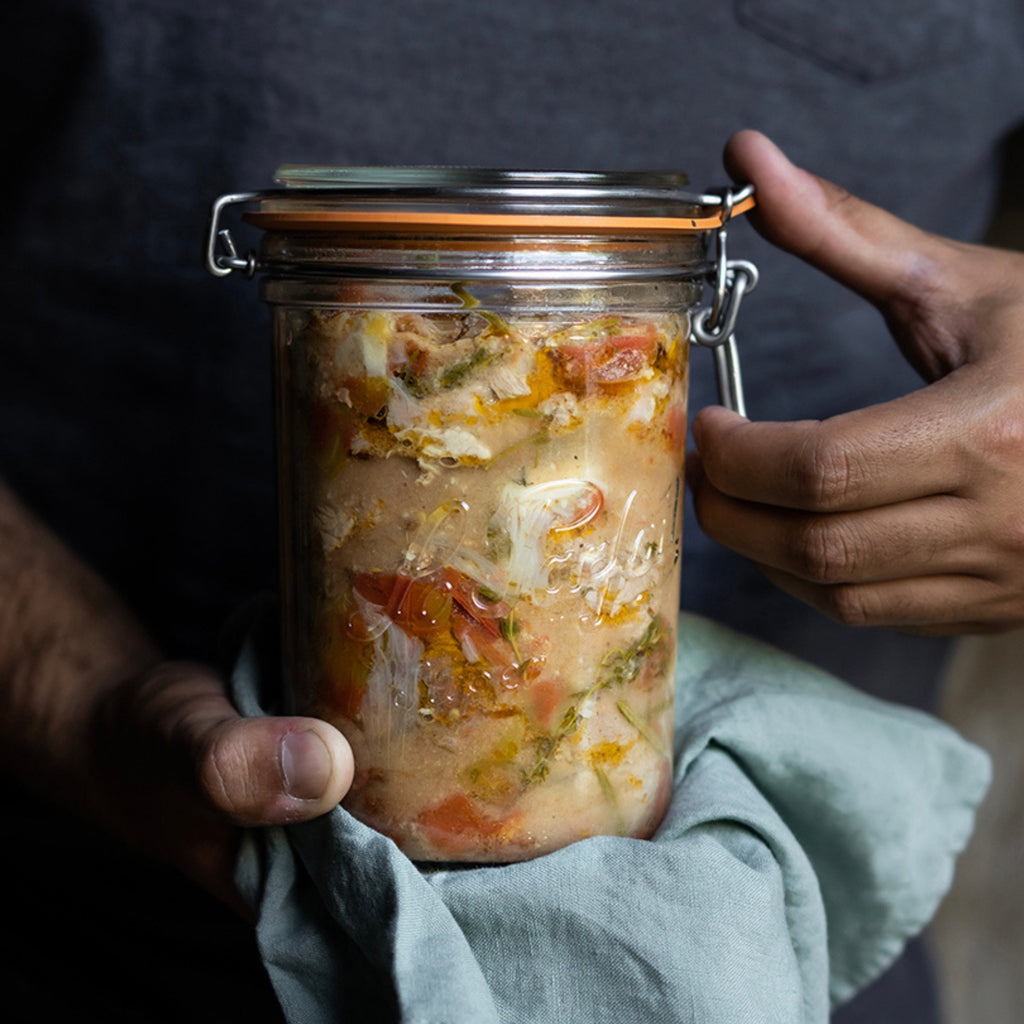 Hands holding a Le Parfait Super Terrine of preserved fish in saffron broth, wrapped in a sage-green cloth.