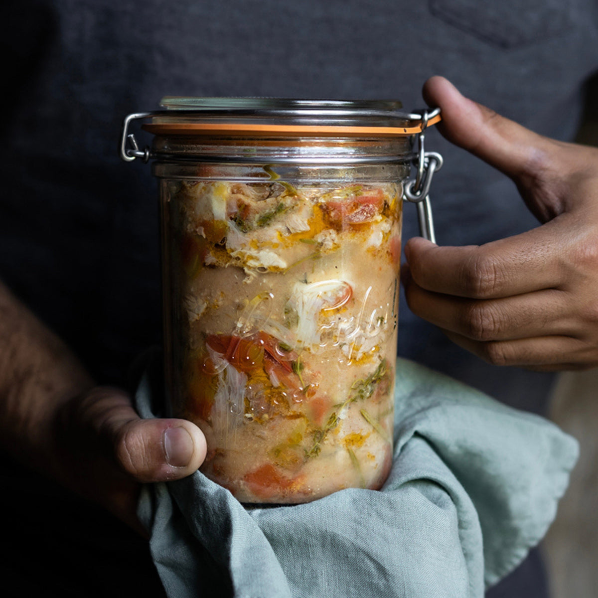 Hands holding a Le Parfait Super Terrine of preserved fish in saffron broth, wrapped in a sage-green cloth.