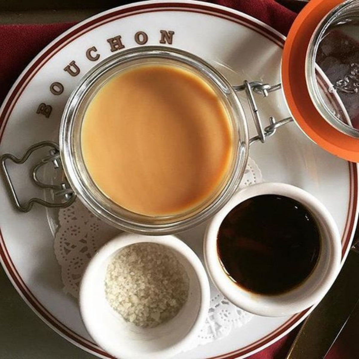 Top-down French bistro plating with a Le Parfait Super Terrine of crème caramel on a Bouchon-branded plate, accompanied by sugar and caramel syrup.