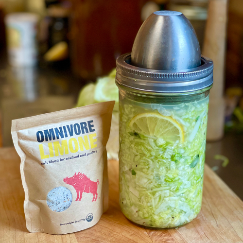 Mason-style fermentation jar of cabbage with ChouAmi fermenting kit and stainless weight, beside an Omnivore Smoke pouch.