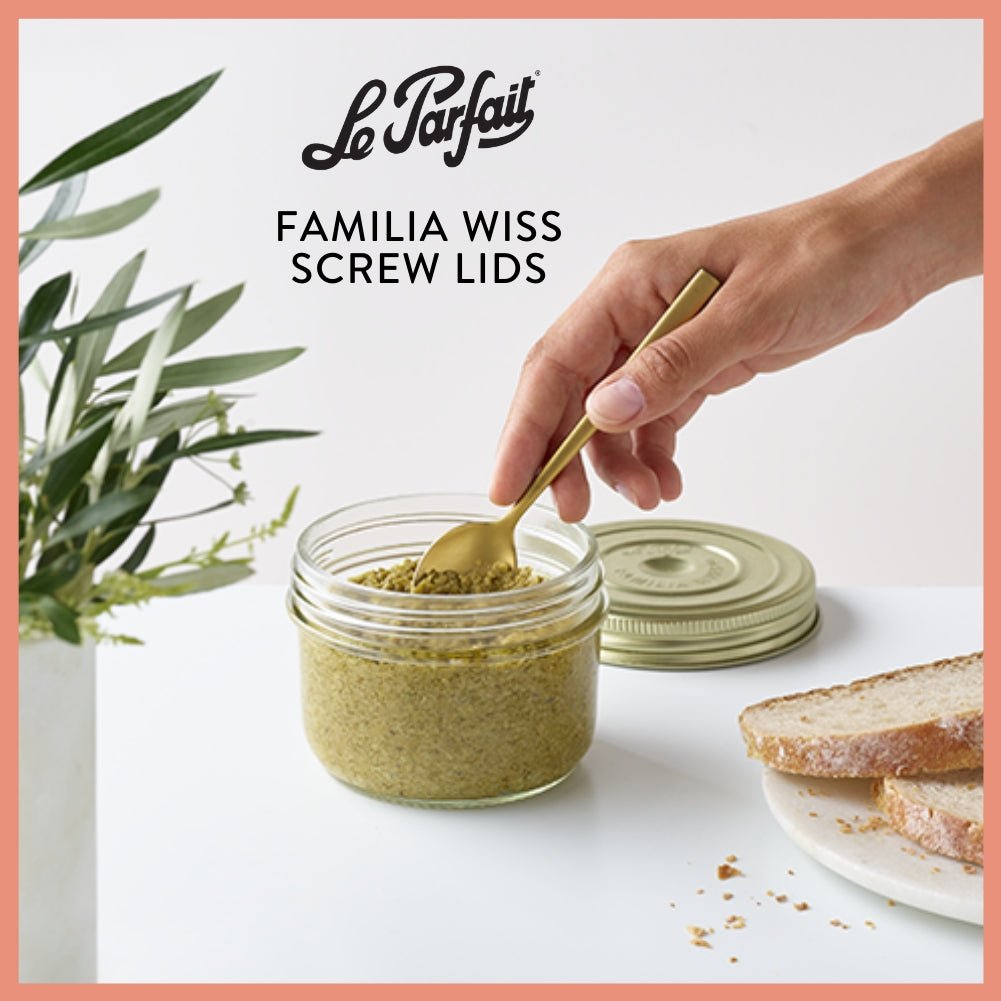 Le Parfait Familia Wiss Screw Lids hero - hand with gold spoon scooping pesto from an open Familia Wiss jar beside sliced bread and olive branches.