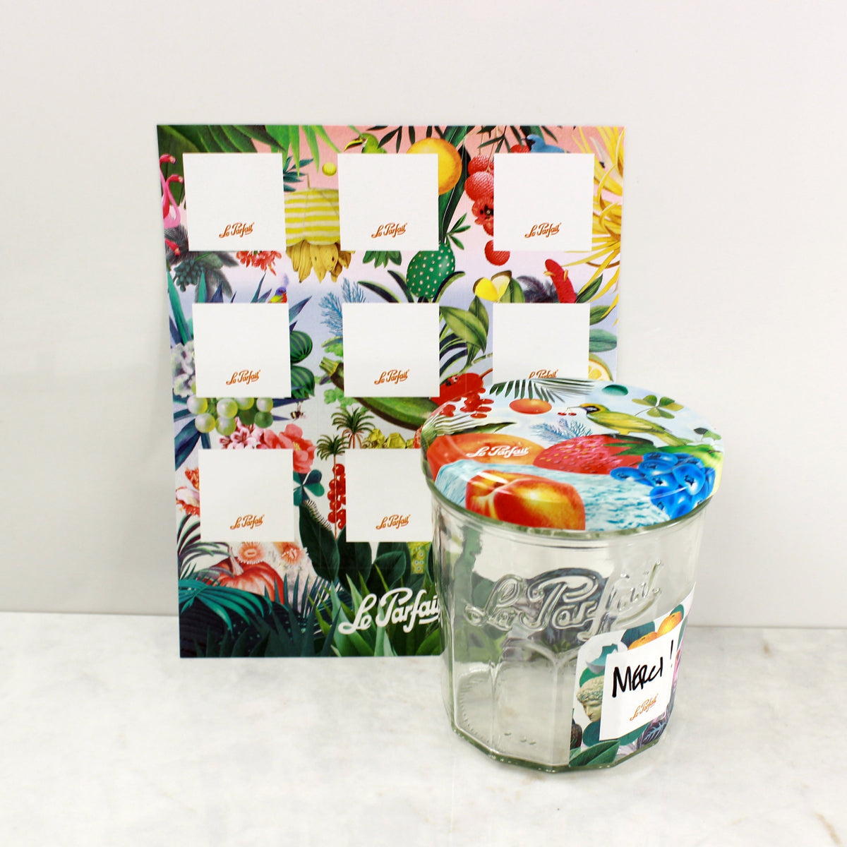Le Parfait Labels tropical jungle sheet beside a Le Parfait jar with matching watercolor-fruit lid and a 'Merci!' handwritten label.
