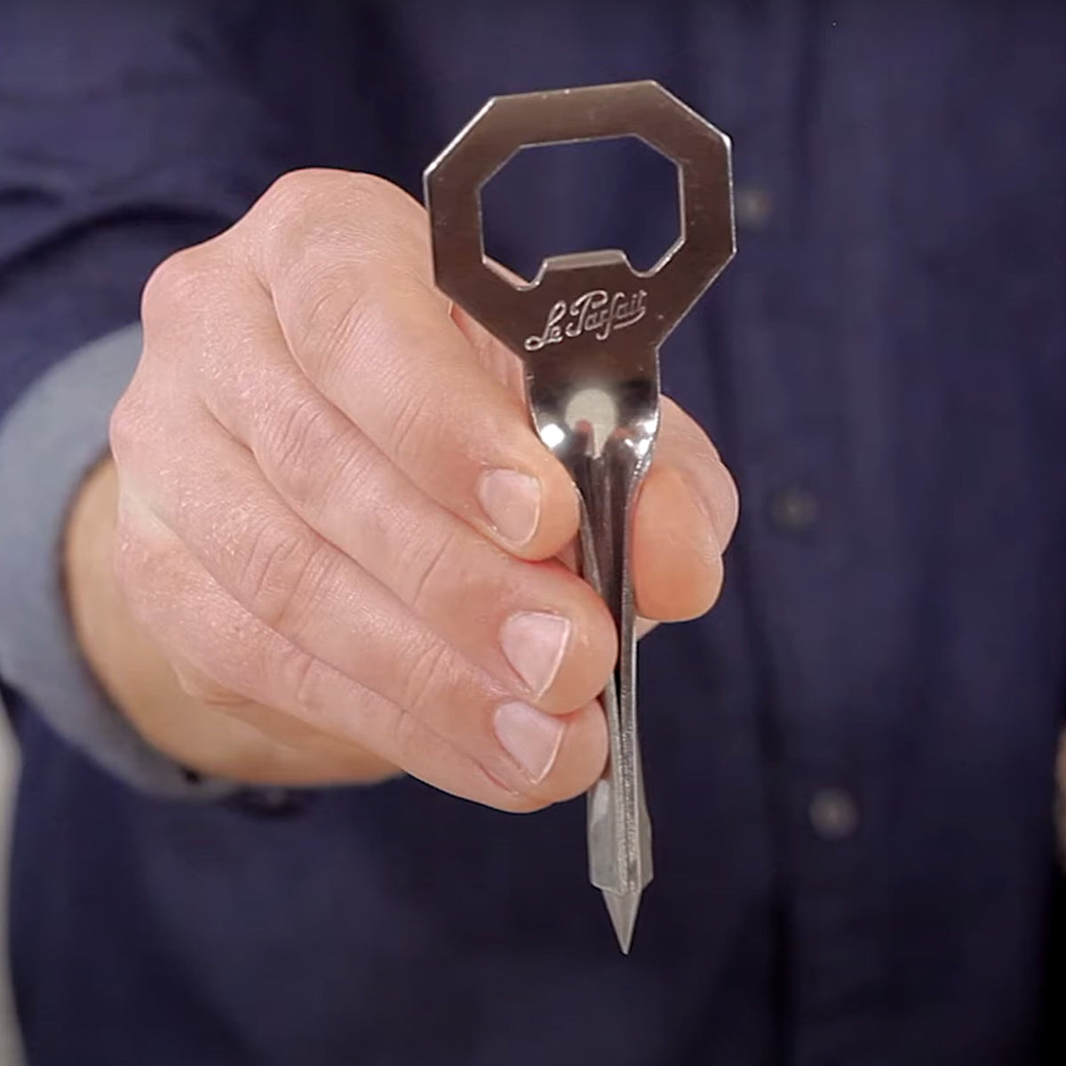 Hand holding a Le Parfait Tire-Rondelle stainless steel jar opener showing the etched Le Parfait branding.