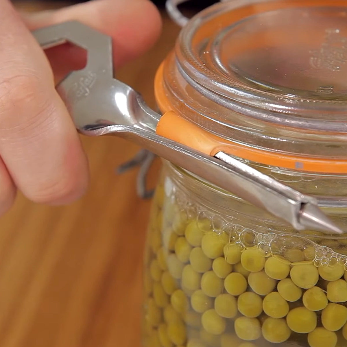Le Parfait Tire-Rondelle opener prying up the orange rubber seal of a clip-top jar of preserved peas.