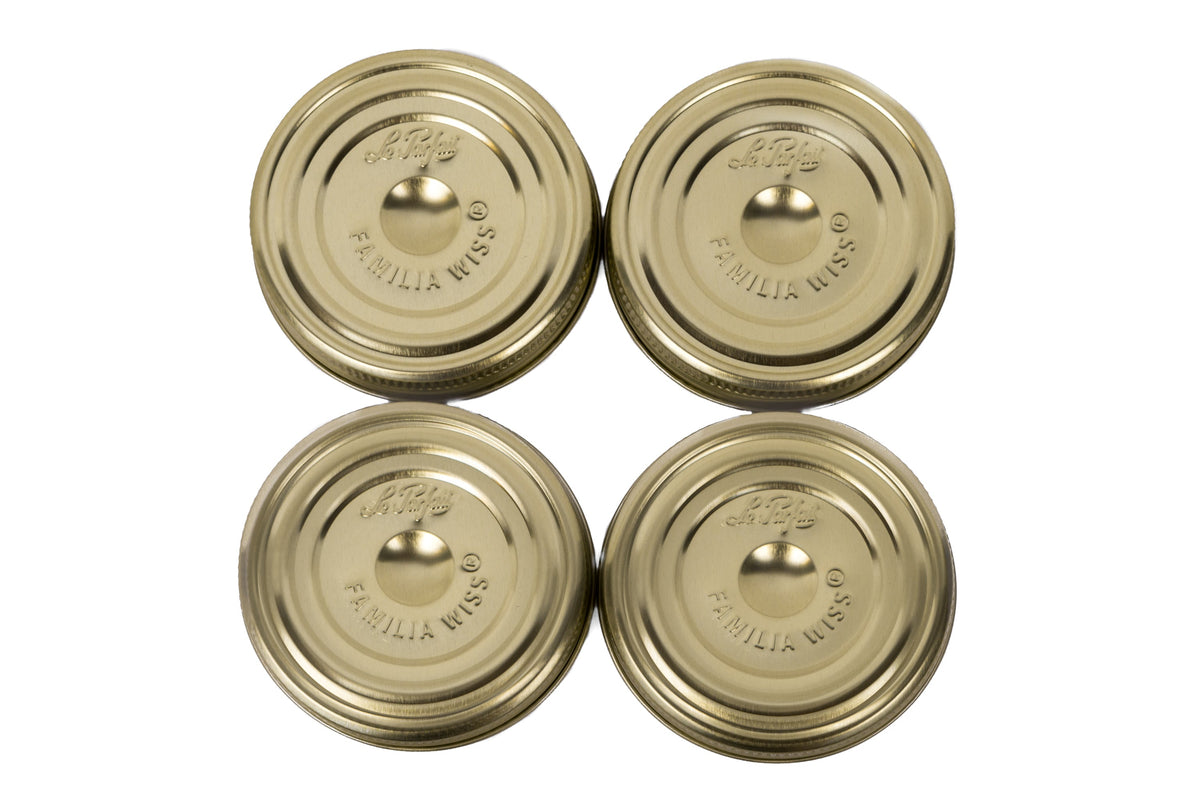 Four Le Parfait gold Familia Wiss metal canning lids with embossed logo, arranged top-down on white.