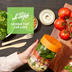 Hand holding a Le Parfait Super Jar of layered quinoa, vegetables, and greens with orange screw lid; tomato vine and canning lid alongside.